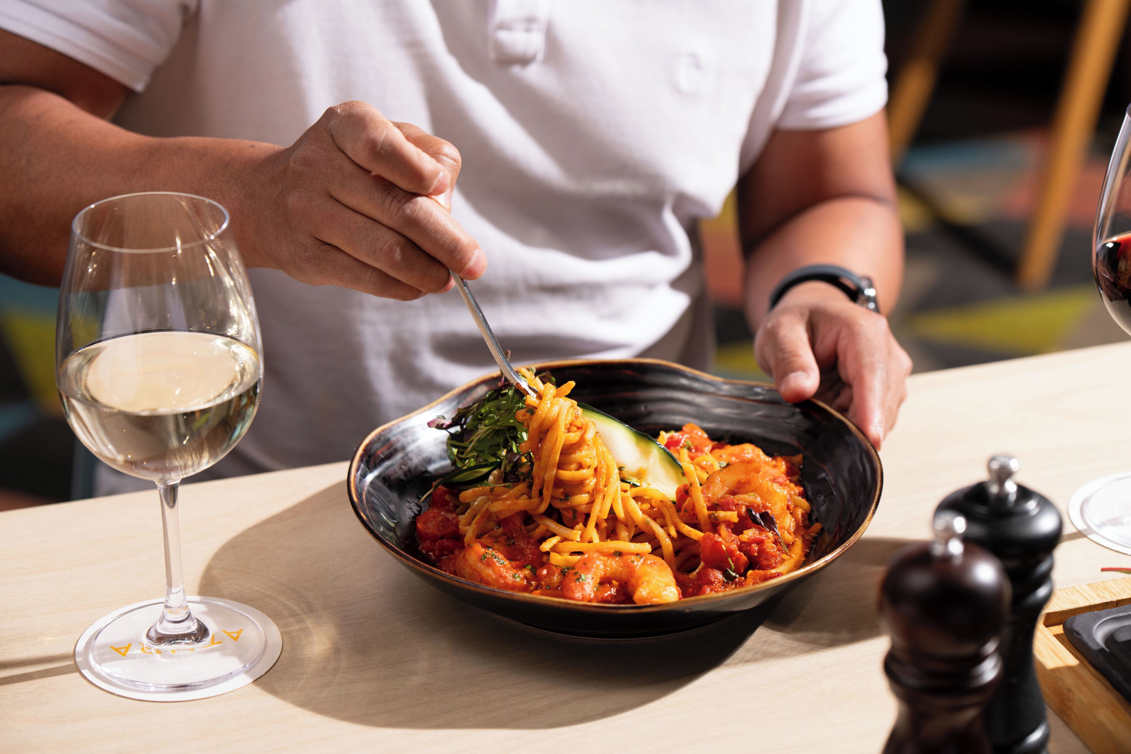 A person dining at a table with a bowl of pasta, a glass of white wine, and a pepper mill.