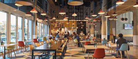 Une cafétéria spacieuse avec de longues tables communes, des chaises colorées et des luminaires suspendus.
