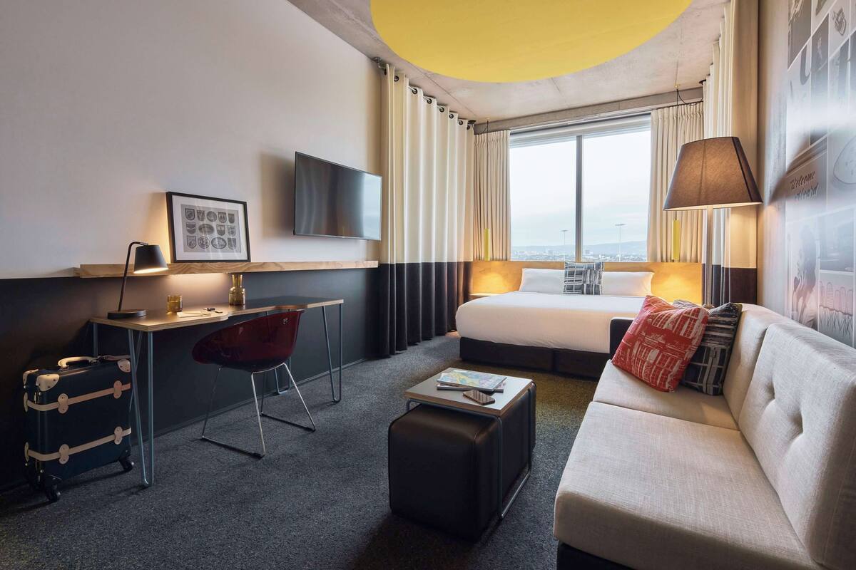 A hotel room with a bed, a desk, a chair, a sofa, a TV, and a window with curtains.