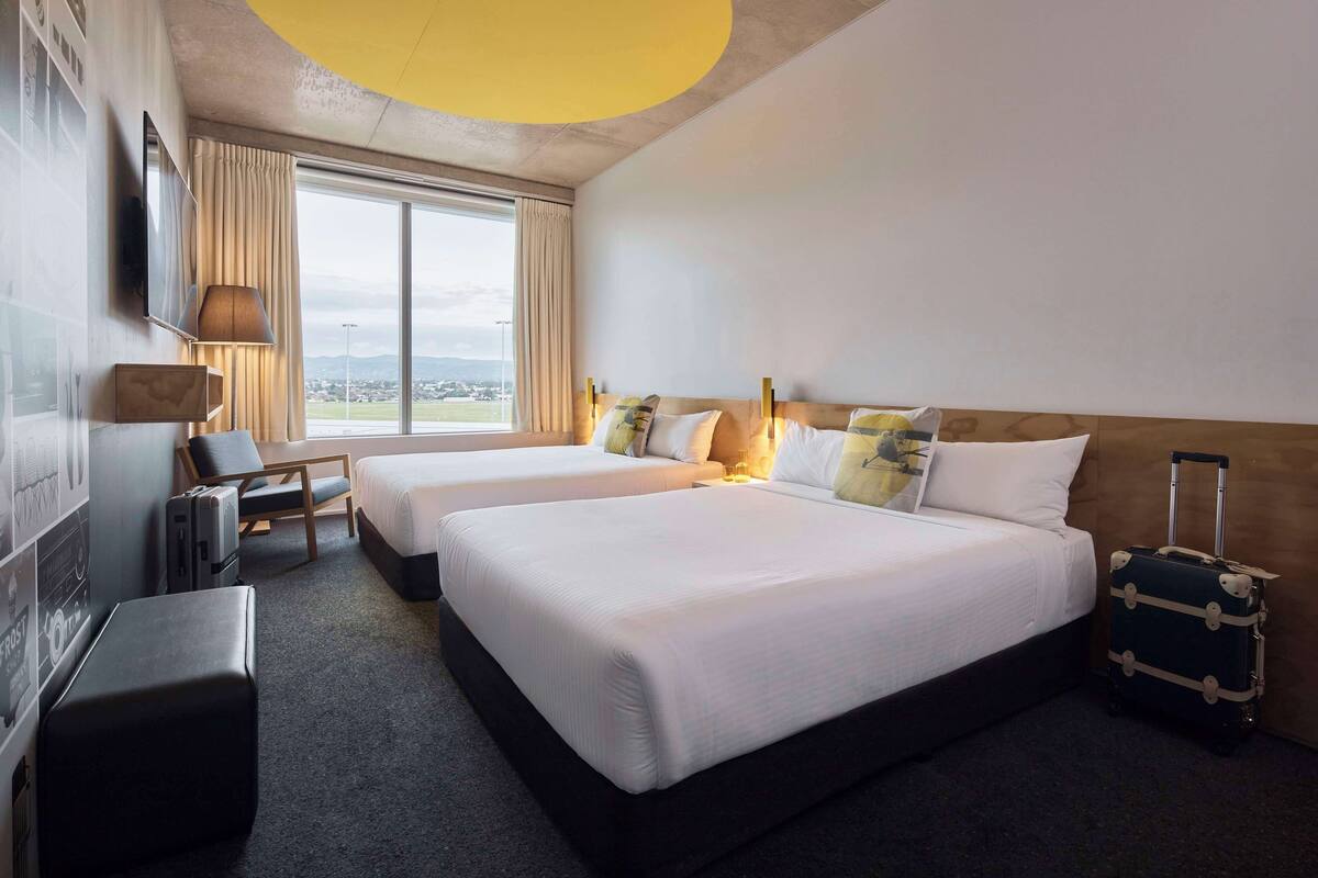 A hotel room with two beds, a desk, a chair, a suitcase, and a large window offering a view.