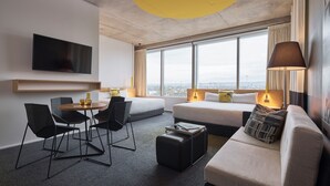 A modern hotel room with a large window, a flat-screen TV, a sofa, a dining table with chairs, and a small ottoman. - Atura Adelaide Airport (Adelaide Airport)