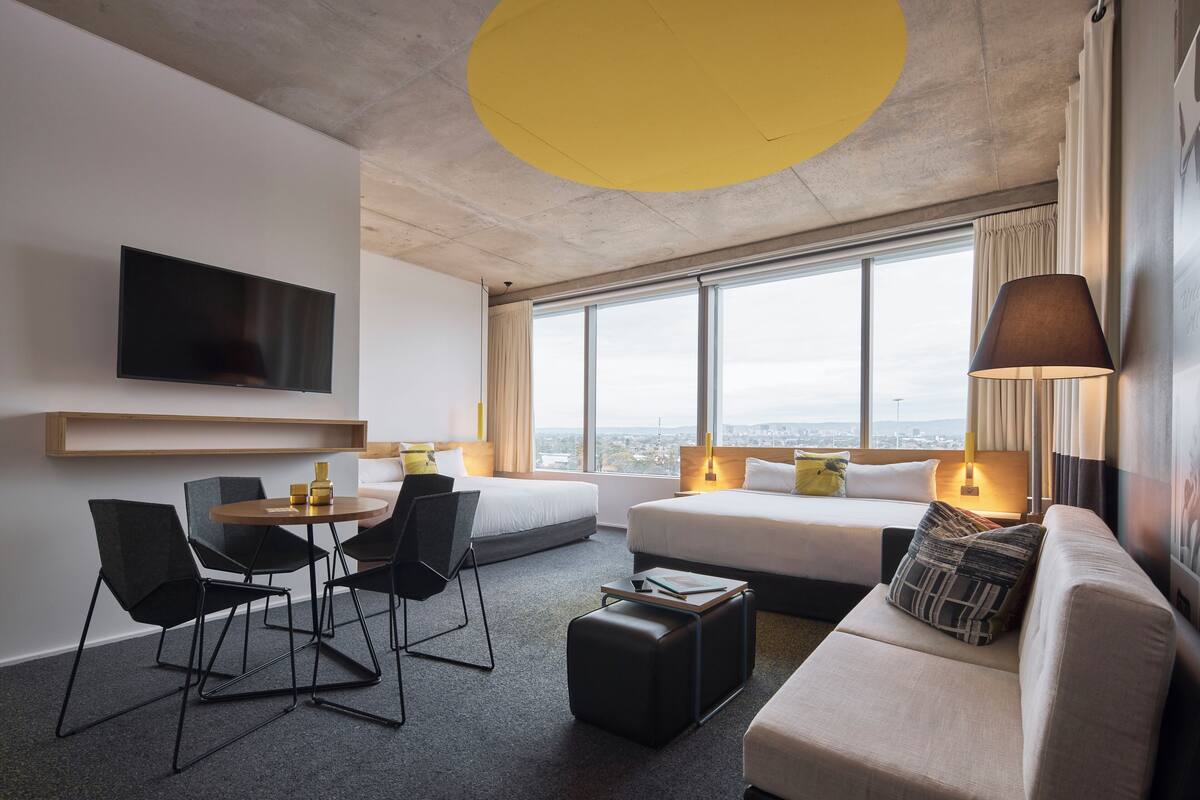 A modern hotel room with a large window, a flat-screen TV, a sofa, a dining table with chairs, and a small ottoman.