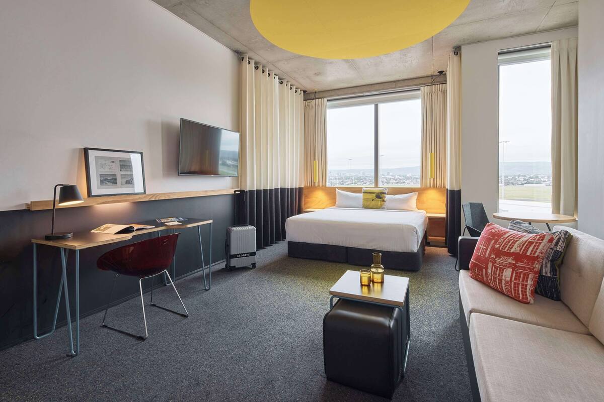 A hotel room with a bed, a desk, a chair, a sofa, a TV, and a view of the city.
