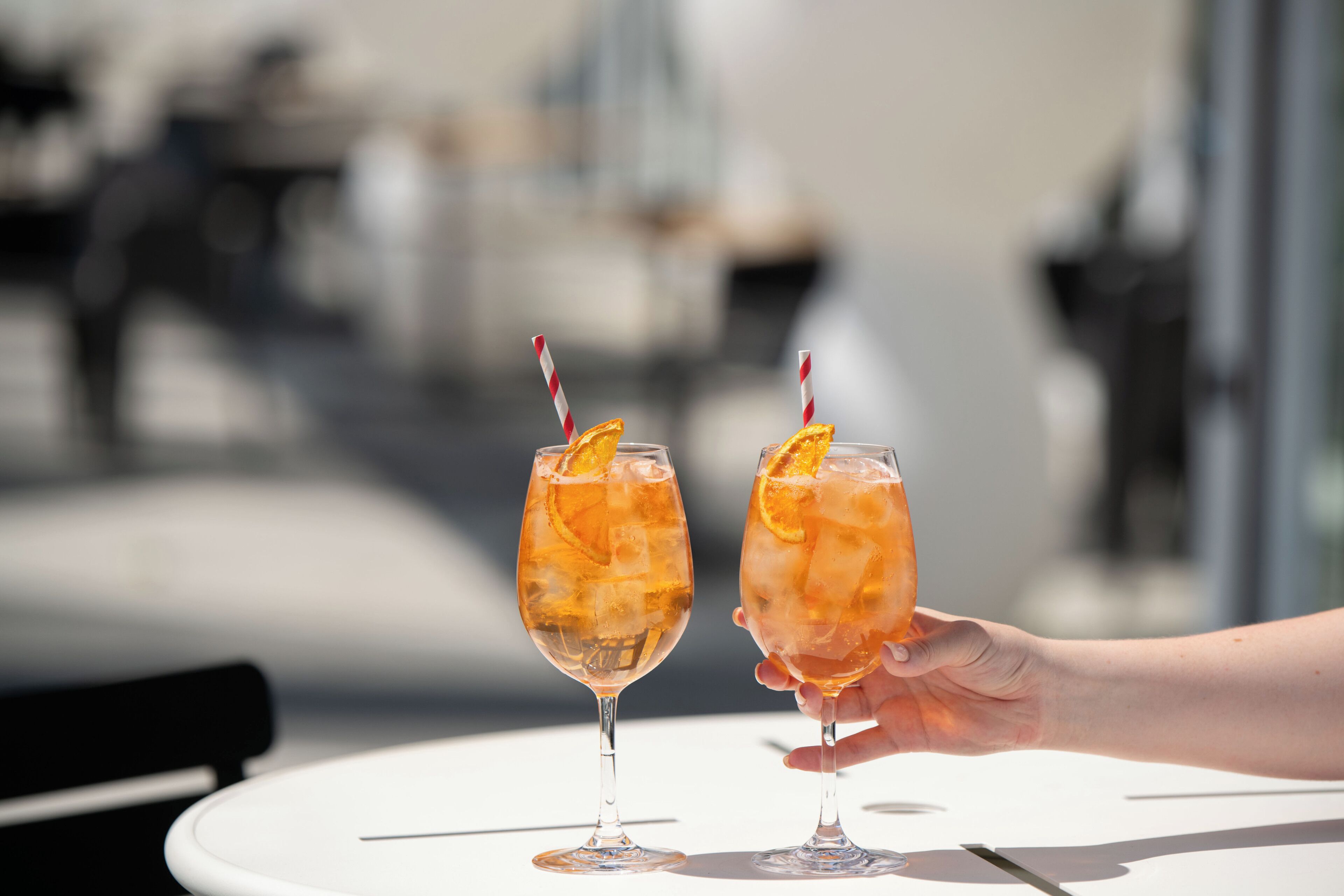Two glasses of a beverage with a slice of orange and a striped straw, held by a hand.