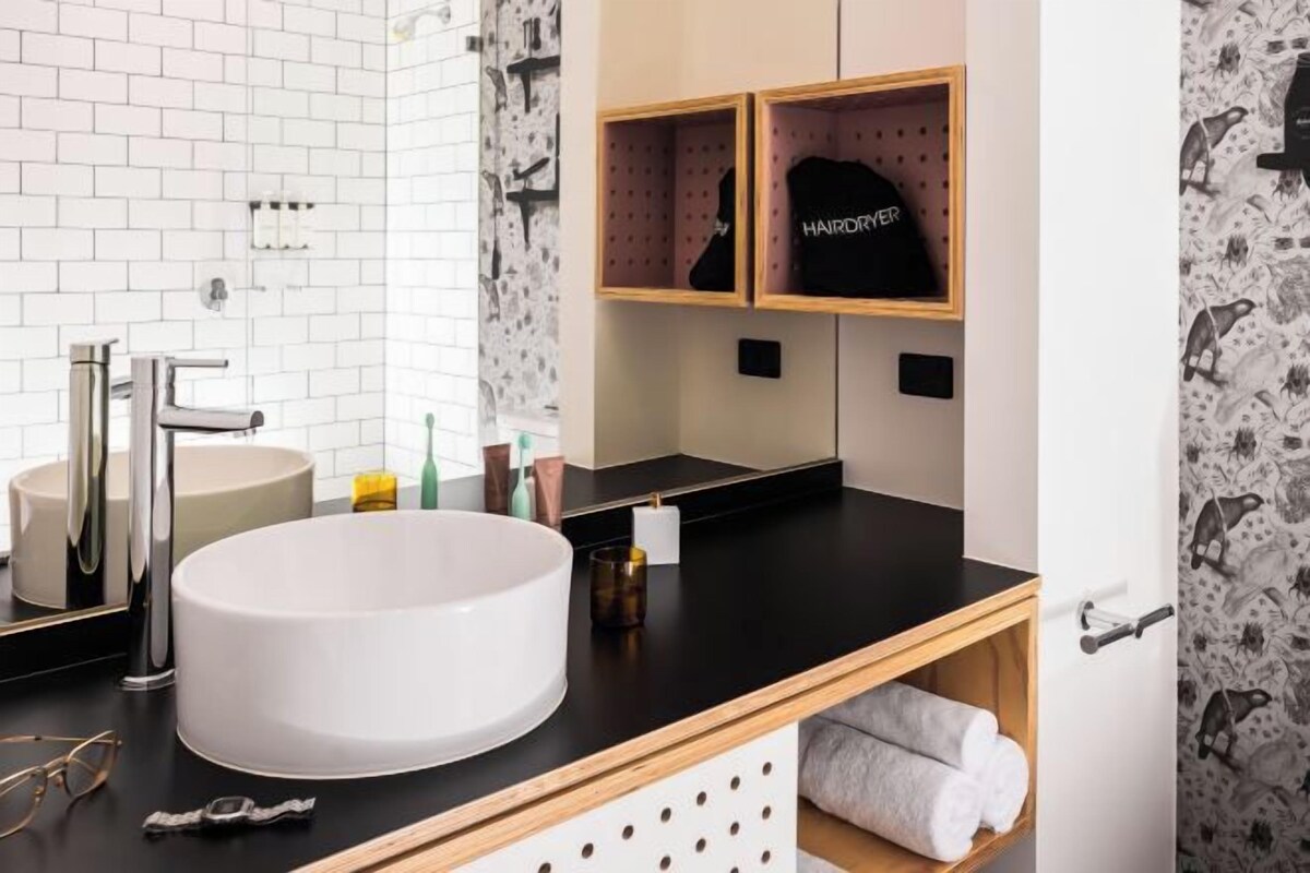 A modern bathroom with a white sink, chrome faucet, and wooden shelves with towels and a black bag labeled "HAIRDRYER".