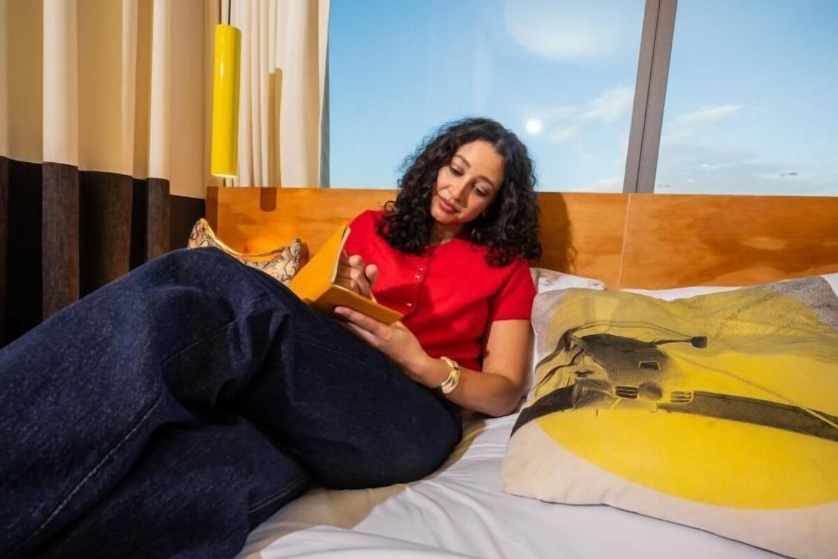 A person lying on a bed in a room with a large window, reading a book.