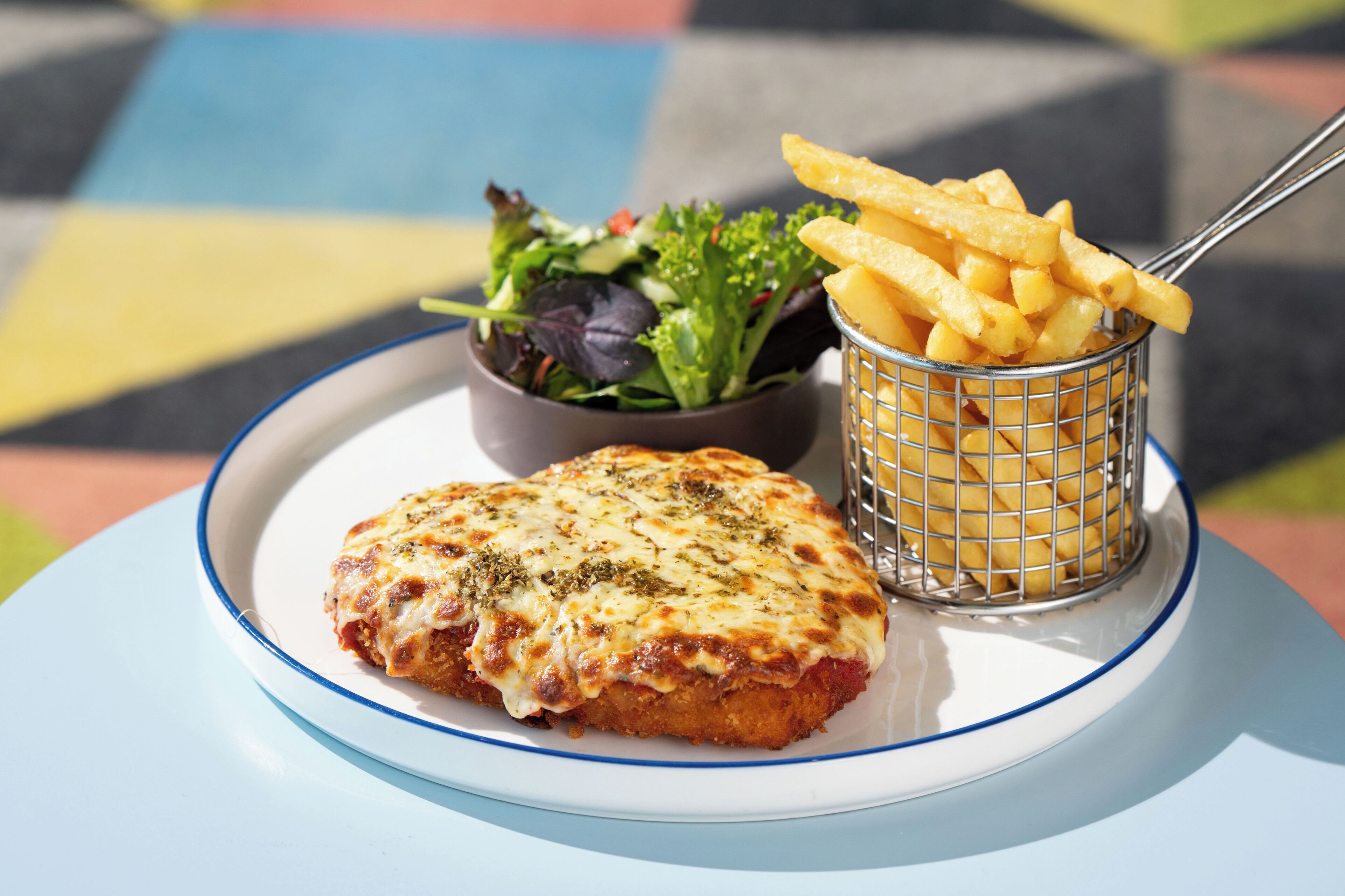 A plate with a slice of pizza topped with cheese and herbs, a small bowl of salad, and a basket of French fries.