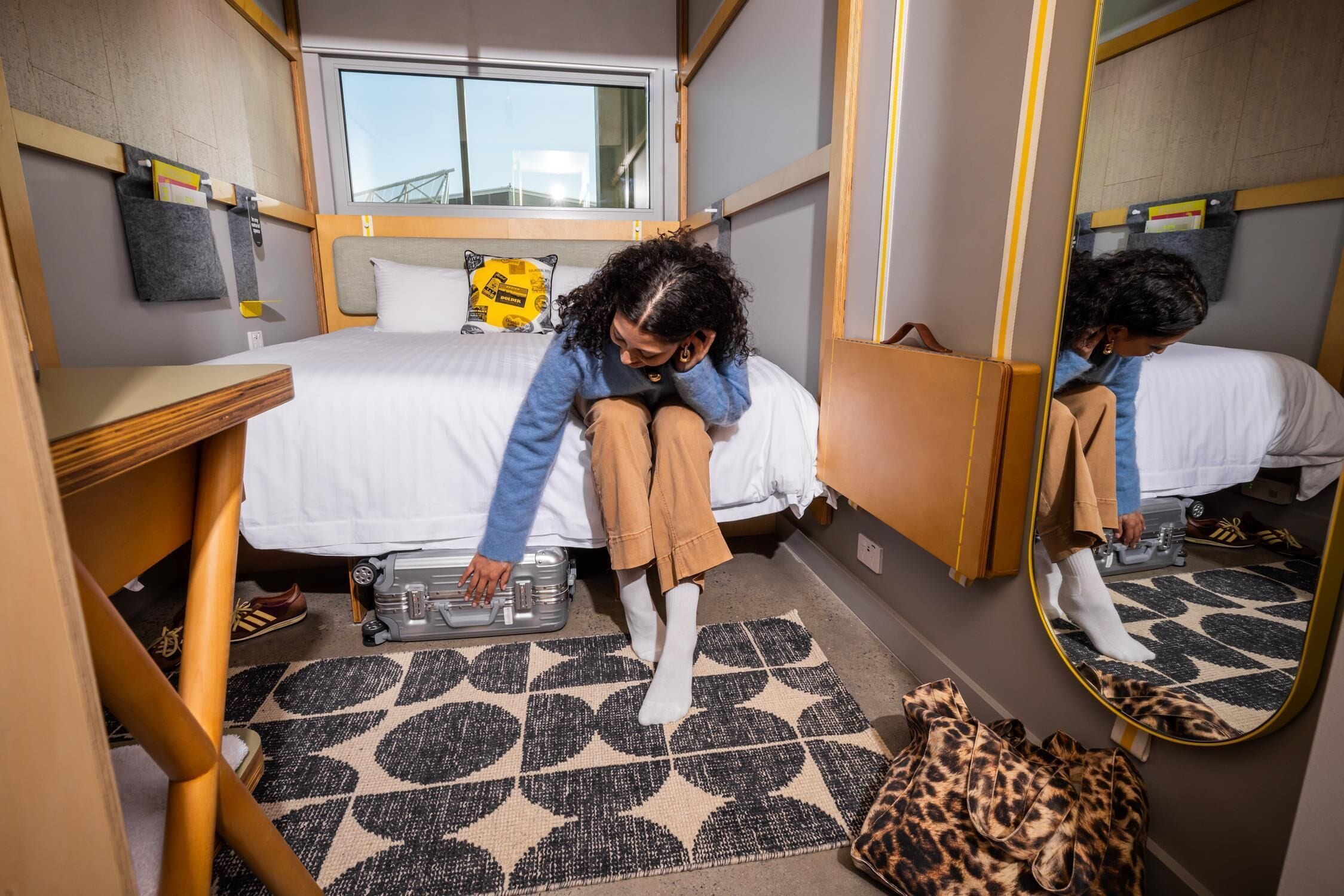 Un dormitorio pequeño con cama, una maleta, un bolso estampado de leopardo y un espejo que refleja la habitación.