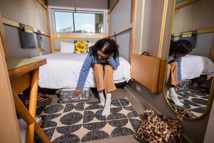 A compact bedroom with a bed, a suitcase, a leopard print bag, and a mirror reflecting the room.