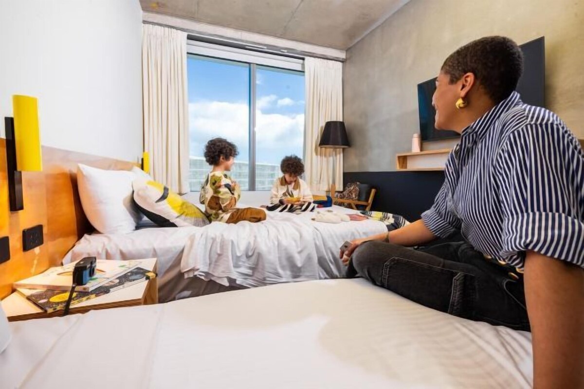 Une chambre d’hôtel avec un lit, une fenêtre avec des rideaux, une lampe et un téléviseur fixé au mur.