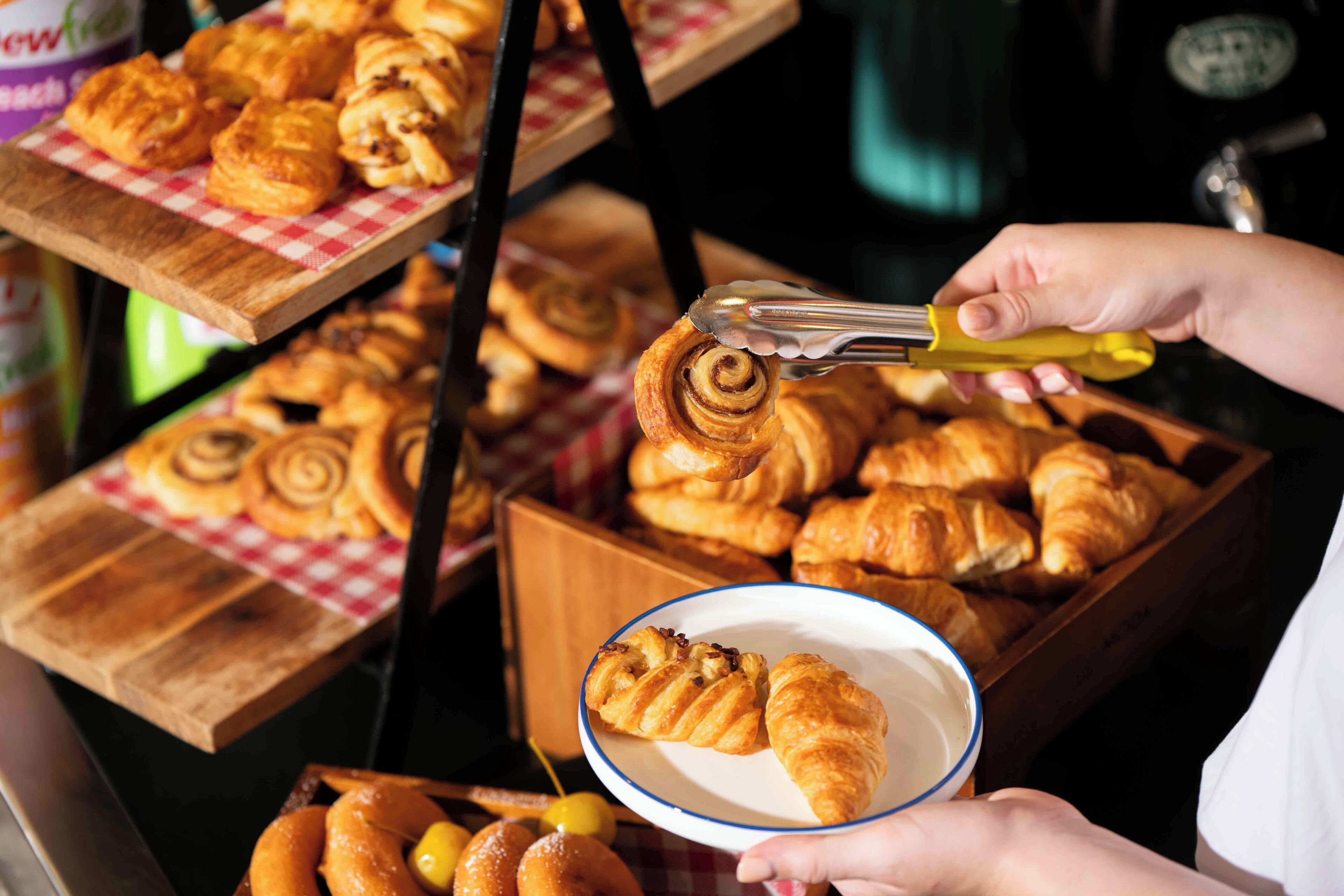 Una persona sosteniendo un plato con un croissant y usando unas pinzas para tomar otra bollería.
