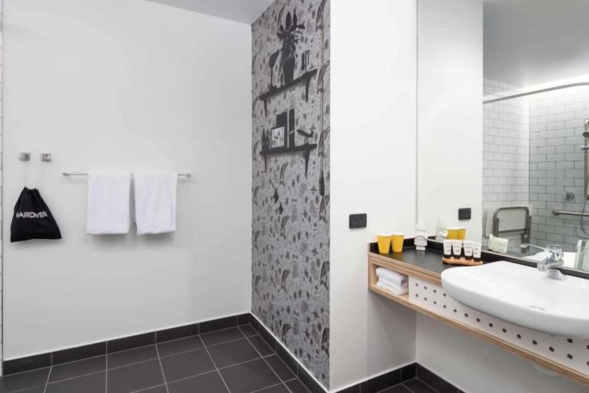 A modern bathroom with a large mirror, a sink, and a shower area. There are towels hanging on a rack and toiletries on a shelf.