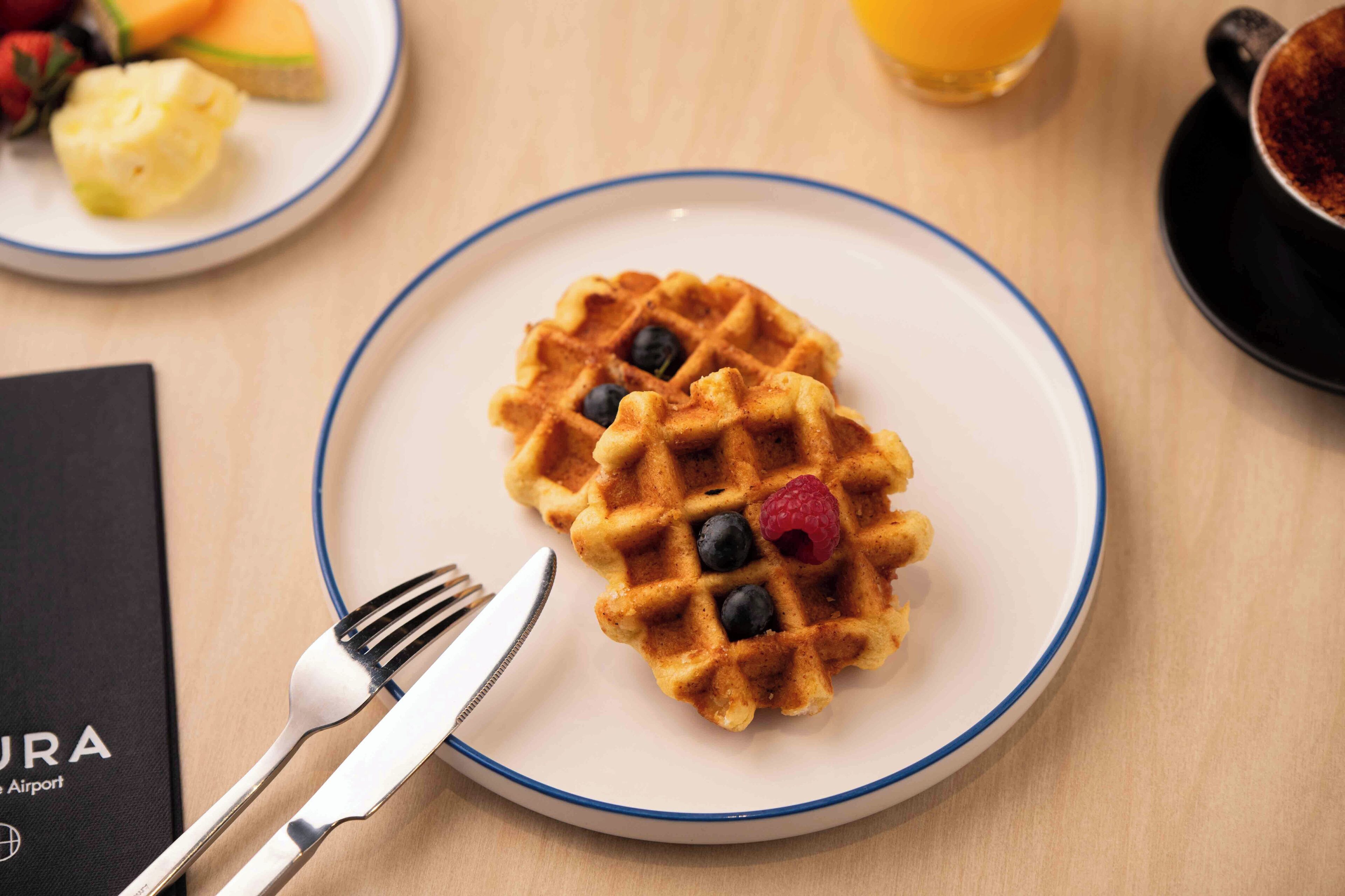 En morgenmadstallerken med en vaffel, blåbær og en hindbær, samt en gaffel og kniv på et bord med en menu.