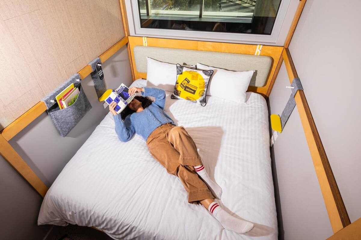 A person lying on a bed with a window above, reading a book.