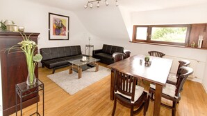A living room with a black sofa, a glass coffee table, a wooden dining table with chairs, a white rug, and a window with a view of greenery. - Ferienwohnung BECKENDORF (Losheim am See)