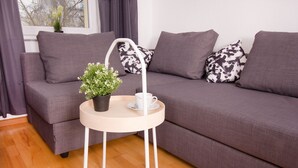 A modern living room with a grey sofa, a small round table with a plant and a cup, and a window with curtains. - Ferienwohnung BECKENDORF (Losheim am See)
