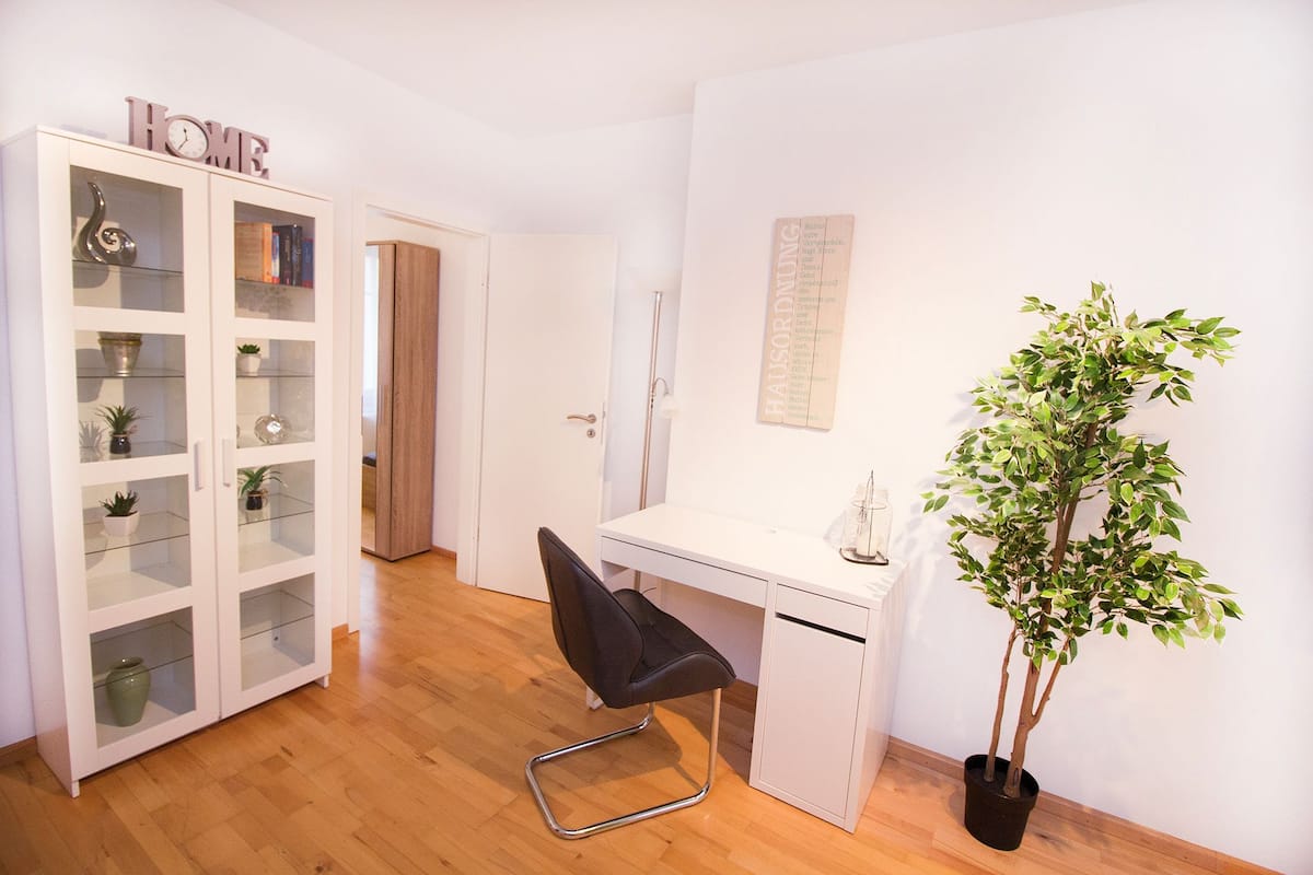A room with a white desk, a black chair, a potted plant, and a cabinet with glass doors.