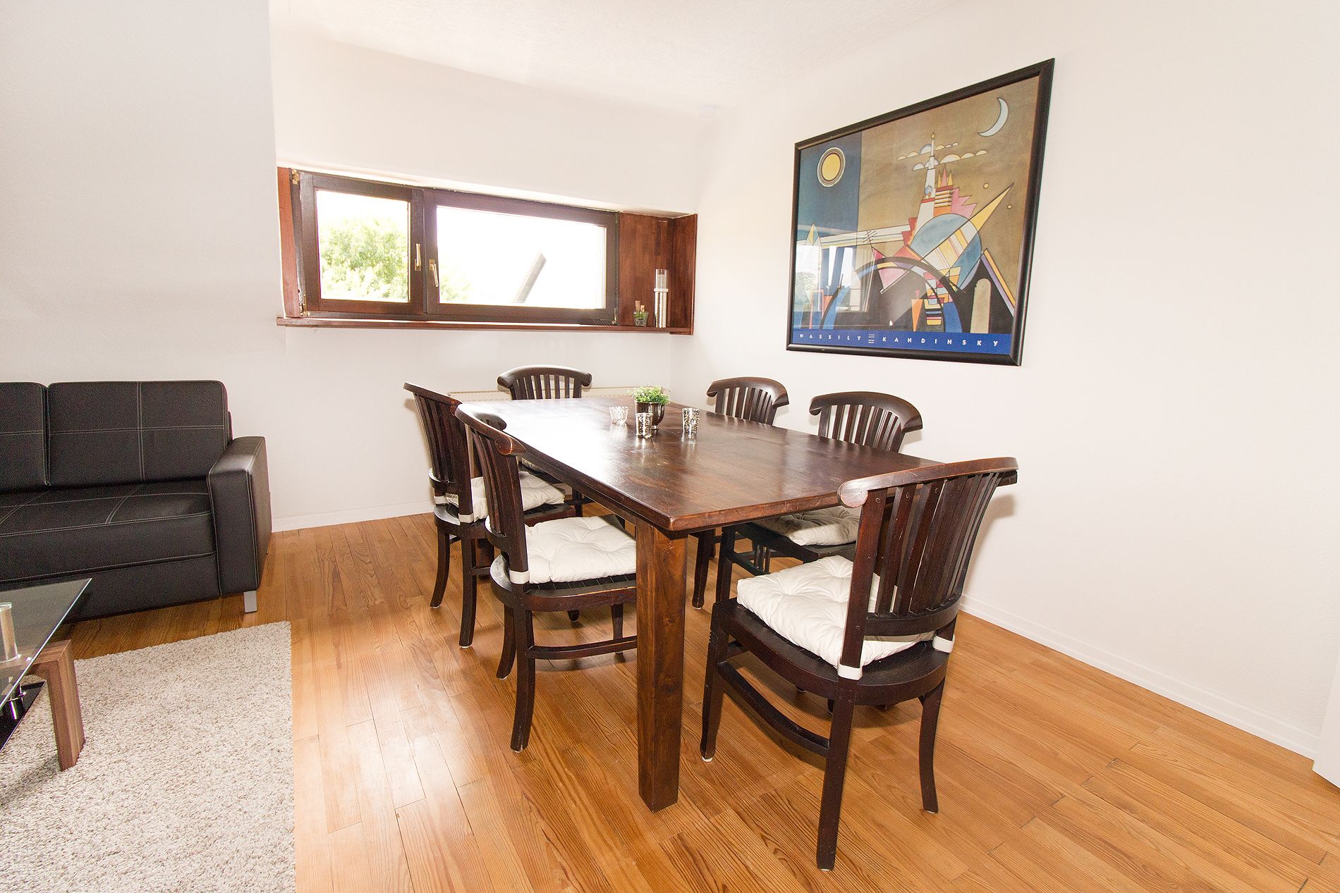 Un área de comedor con una mesa de madera y seis sillas, un sofá negro y un cuadro enmarcado en la pared.