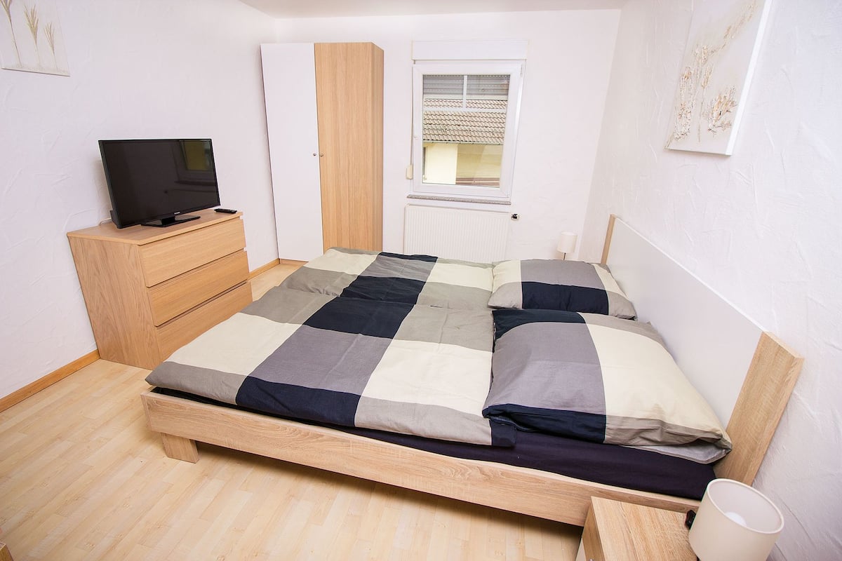 A bedroom with a wooden bed, a TV on a wooden dresser, and a window with a view of a tiled roof.