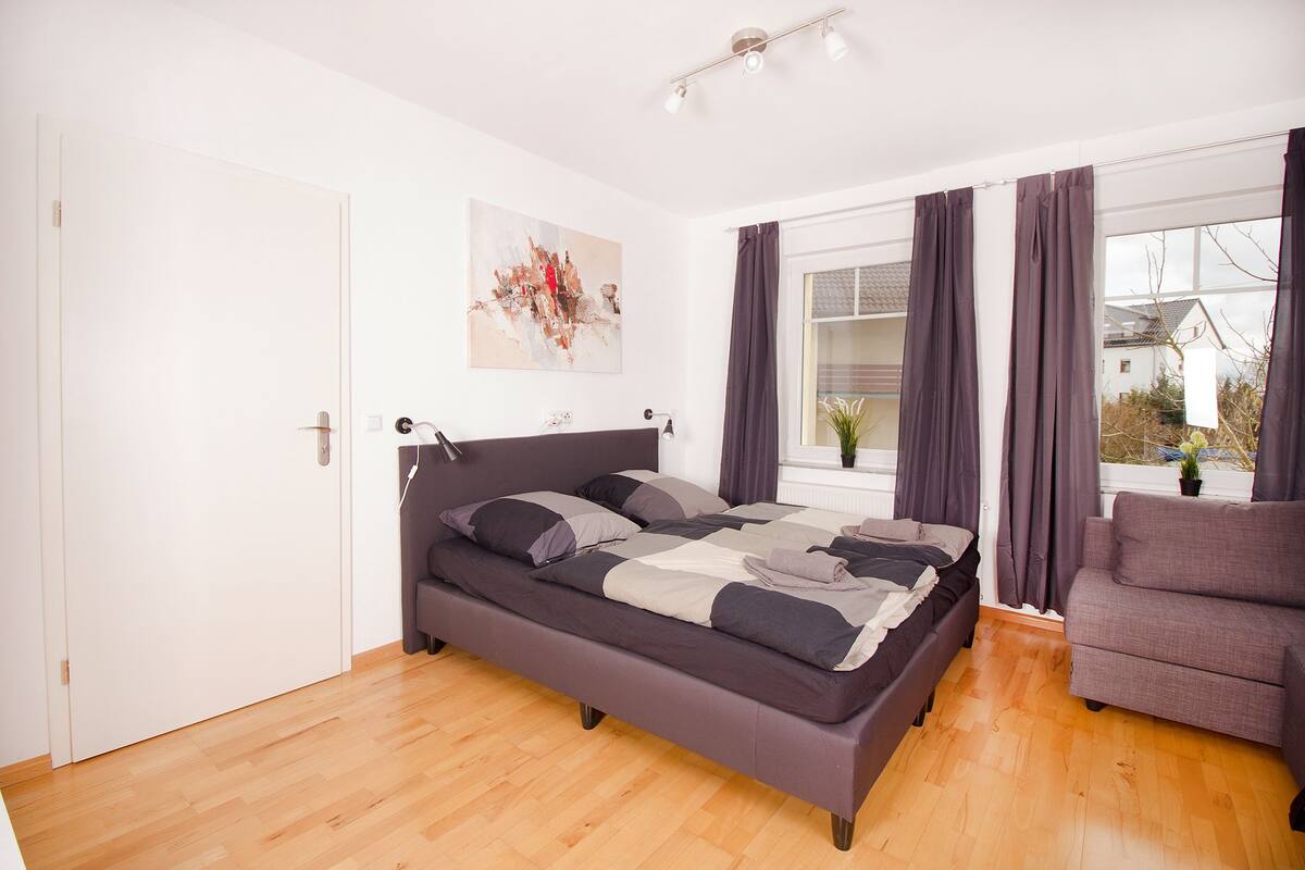 A modern bedroom with a large bed, a chair, a window with curtains, and a painting on the wall.