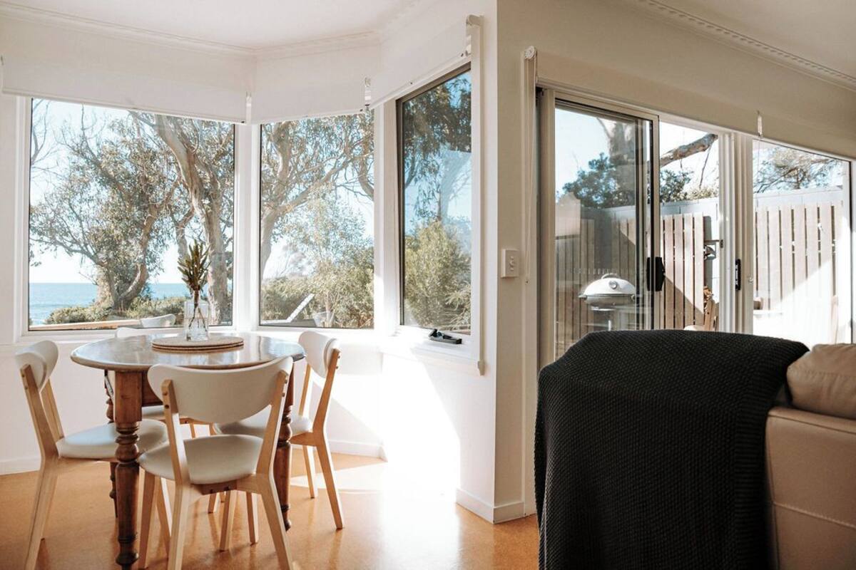 A dining area with a round table and chairs, large windows offering a view of trees and a body of water, and a sliding glass door leading to an outdoor area.