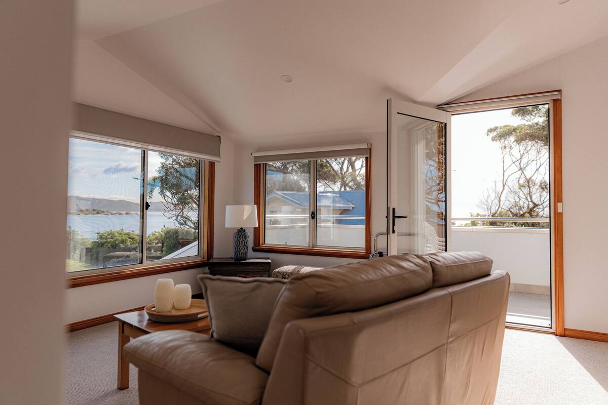 A modern living room with a sofa, a small table with candles, and large windows offering a view of the sea.