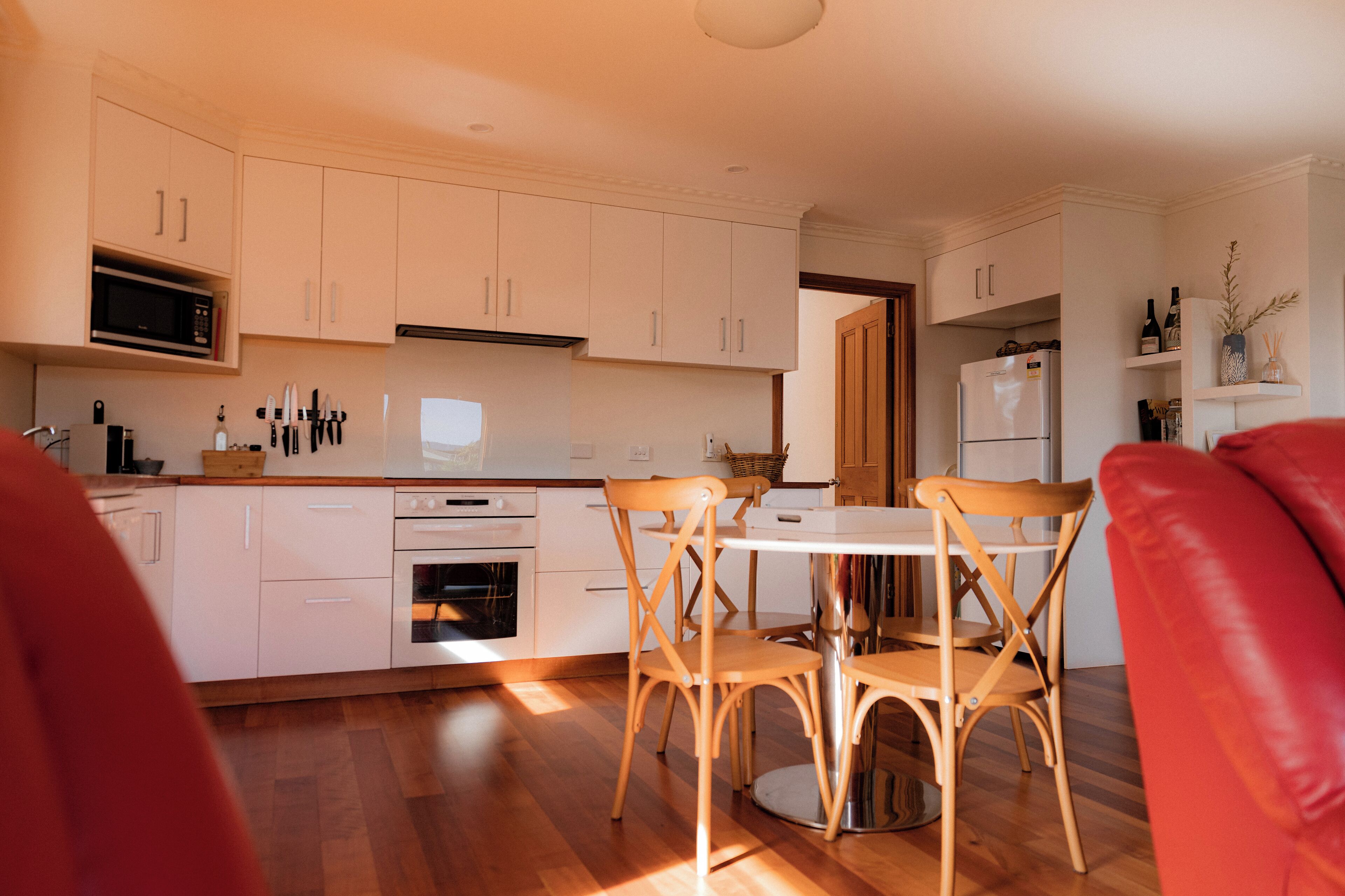 A modern kitchen with white cabinets, a microwave, and a refrigerator. There is a dining table with chairs and a red couch in the foreground.