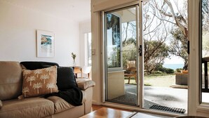 Ein Wohnzimmer mit beiger Couch, einem Holztisch und einer Glastür, die zu einem Außenbereich mit Meerblick und viel Grün führt.