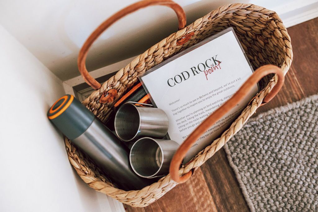 A woven basket containing a metal water bottle, a stainless steel tumbler, and a booklet titled 'COD ROCK POOMPT'.