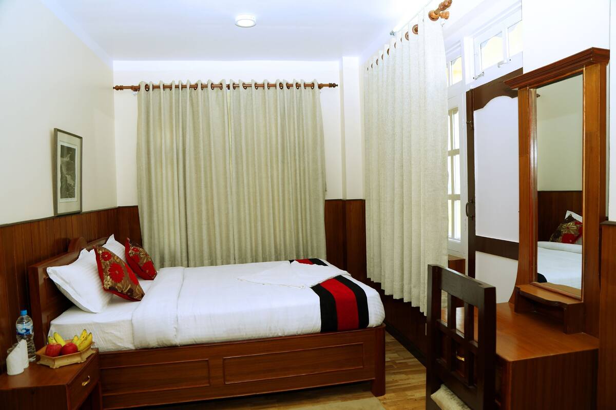 A neatly made bed with a white and red striped blanket, a wooden nightstand with a fruit bowl, a mirror, and a wooden chair.