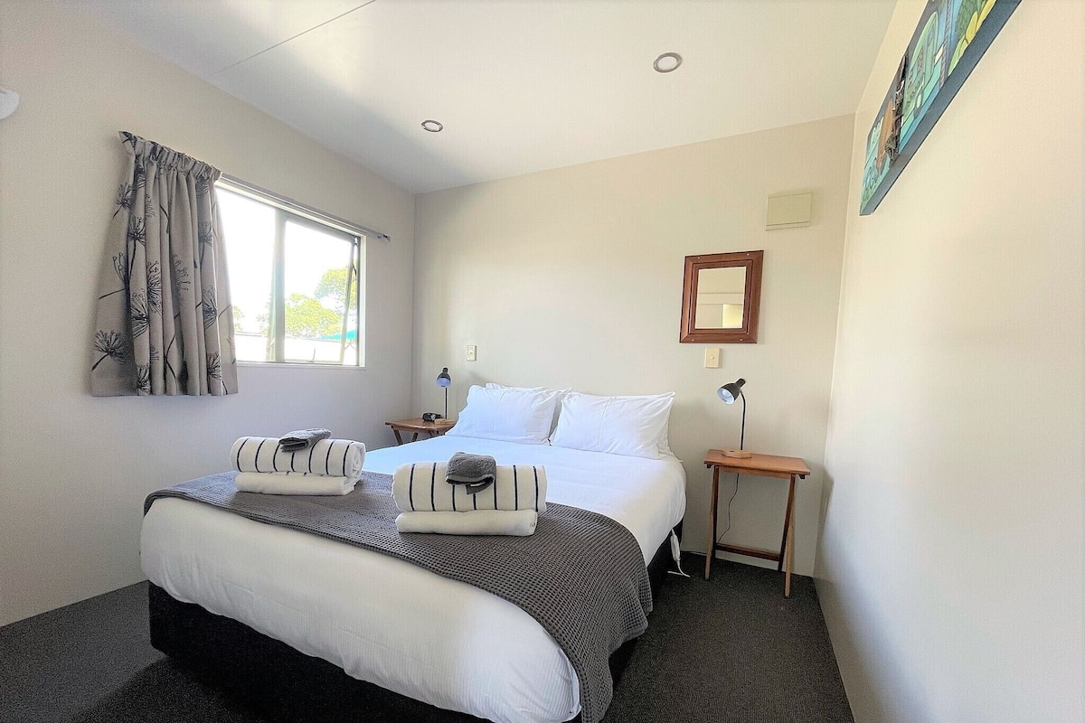 Habitación de hotel con una cama grande, una mesita de noche con lámpara, una ventana con cortinas y un espejo montado en la pared.