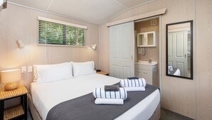 A neatly arranged hotel room with a bed, bedside tables, a window with blinds, a mirror, and a wardrobe. - Athenree Hot Springs & Holiday Park (Waihi Beach)
