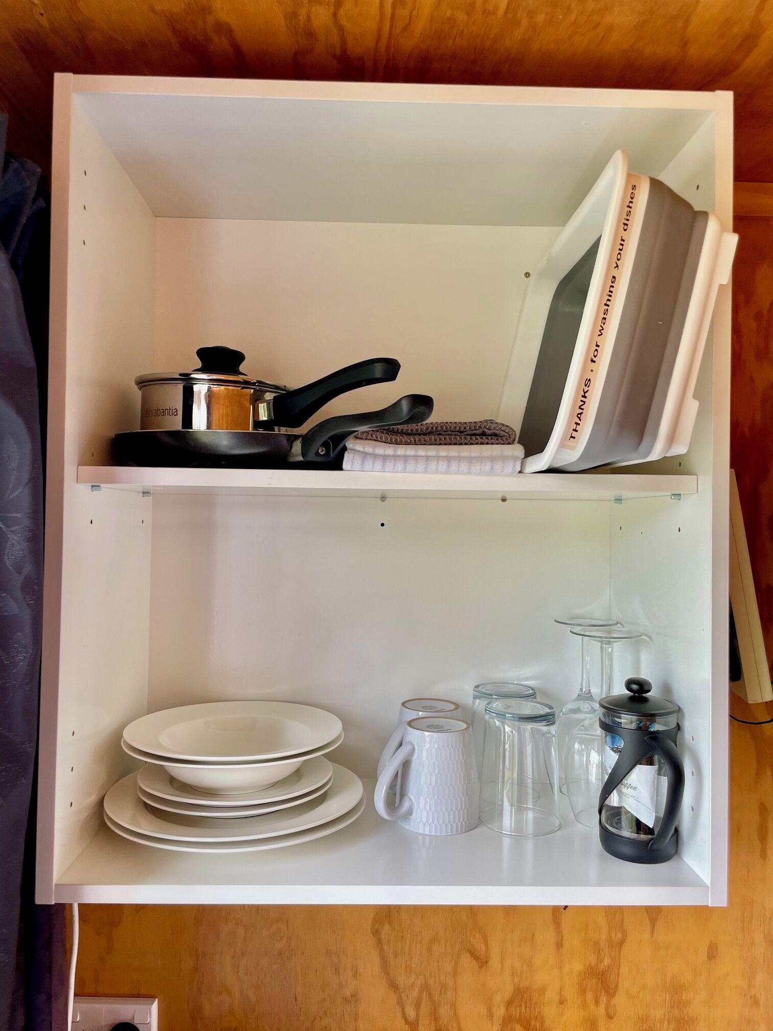 Un estante blanco con utensilios de cocina, incluyendo una olla, sartén y un libro de recetas. Abajo, se apilan platos, una taza blanca y recipientes de vidrio.