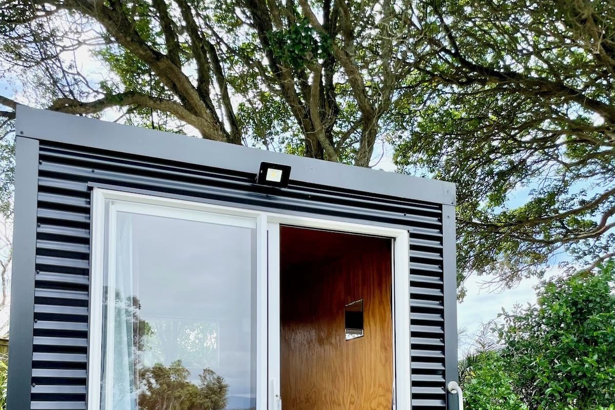 Una estructura moderna y compacta al aire libre con un ventanal de vidrio que da a un interior de madera, una pequeña mesa y dos sillas.