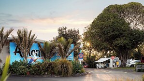 A parking area with a stop sign, a blue building with a sign reading "Agi", and a large tree. - Athenree Hot Springs & Holiday Park (Waihi Beach)