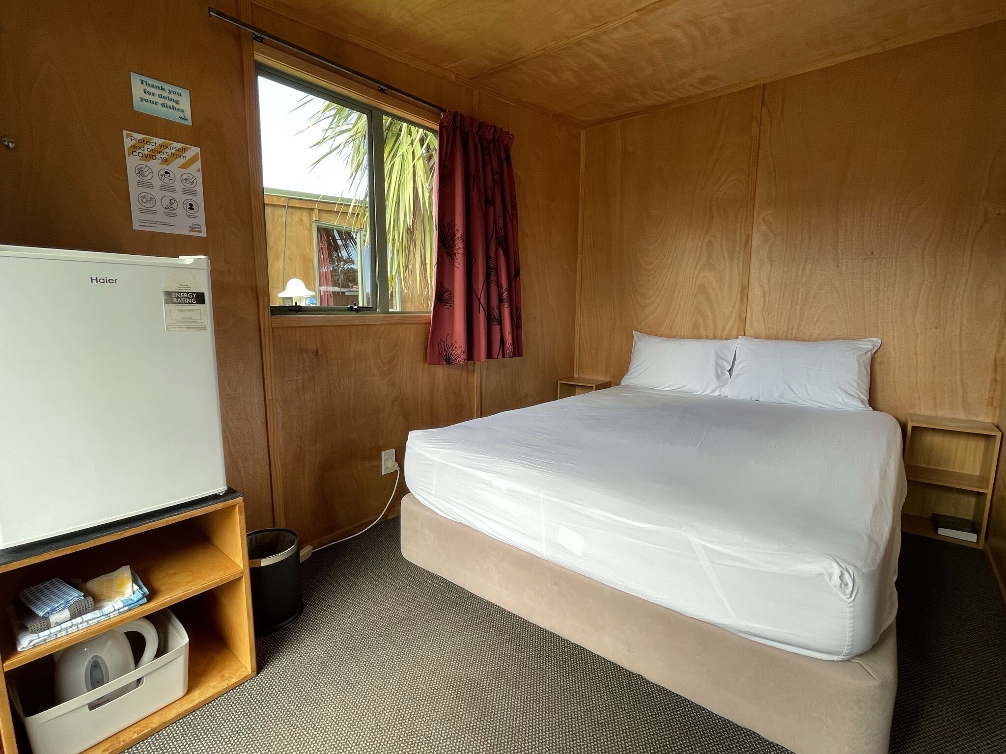 A room with a bed, a white refrigerator, a window with a curtain, and a wooden wall.