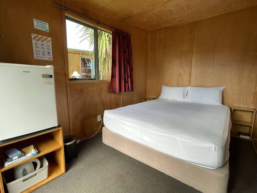 A room with a bed, a white refrigerator, a window with a curtain, and a wooden wall.