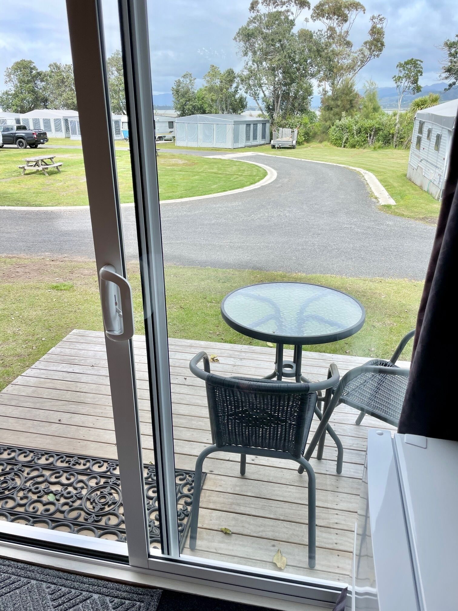 Una terraza con una mesa de vidrio y dos sillas, con vista a un camping con tiendas de campaña y un camino serpenteante.