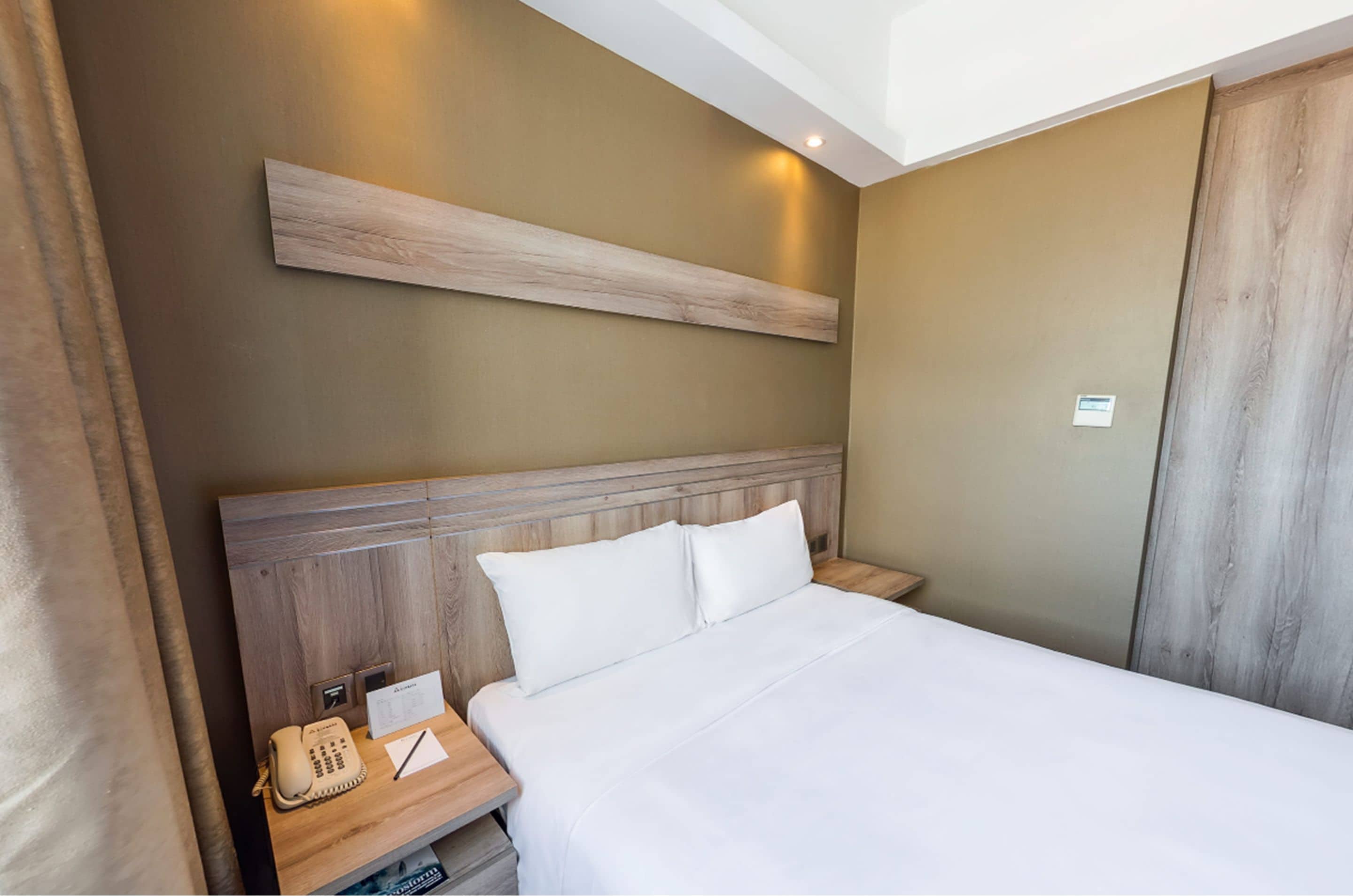 A hotel room with a wooden headboard, a white bedspread, a bedside table with a telephone and a notepad, and a wall-mounted light fixture.