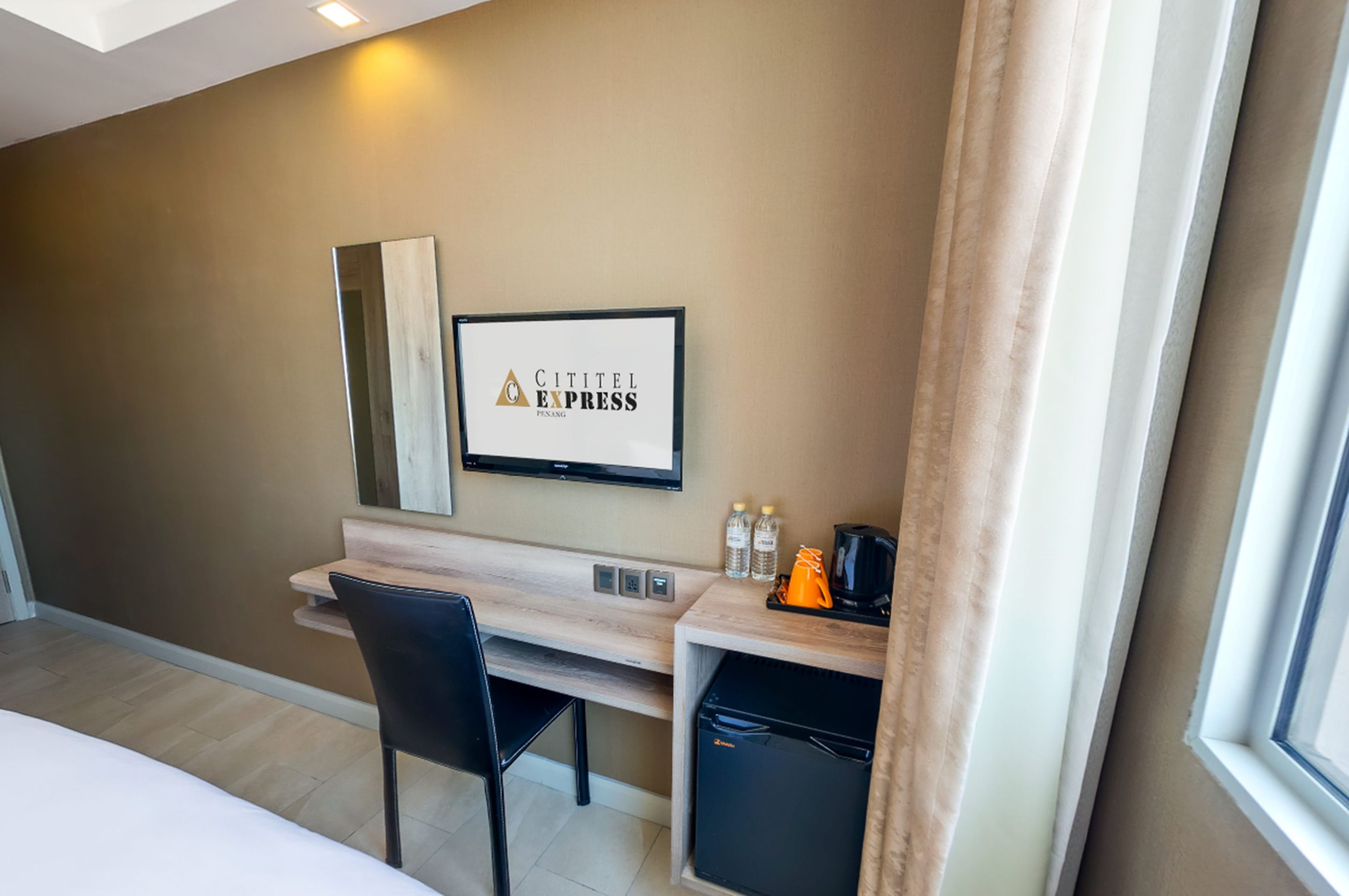 Habitación de hotel con cama, escritorio con silla, refrigerador pequeño, televisor montado en la pared y ventana con cortinas.