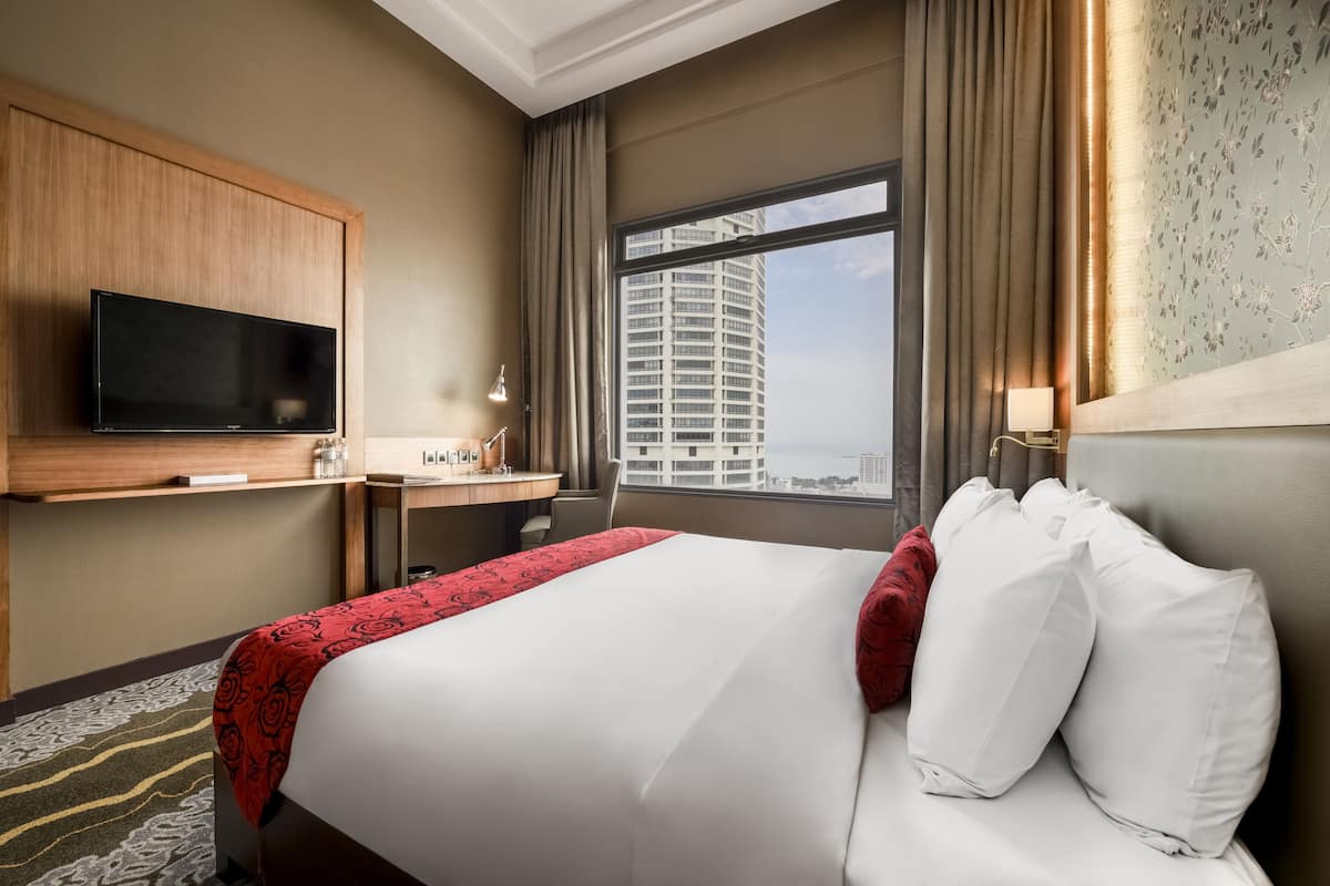 Een hotelkamer met een bed, een aan de muur bevestigde televisie, een bureau met een lamp en uitzicht op een hoog gebouw door het raam.