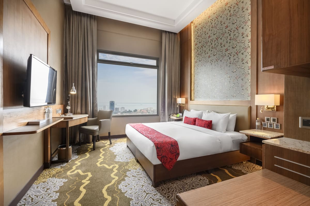Hotelkamer met een groot bed, een bureau met een televisie, een stoel, een bureaulamp, een nachtkastje met een lamp, en uitzicht op de stad door een groot raam.