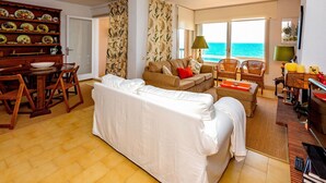 Sala de estar com sofá, mesa de jantar, cadeiras, estante de livros e vista para o mar através de grandes janelas.