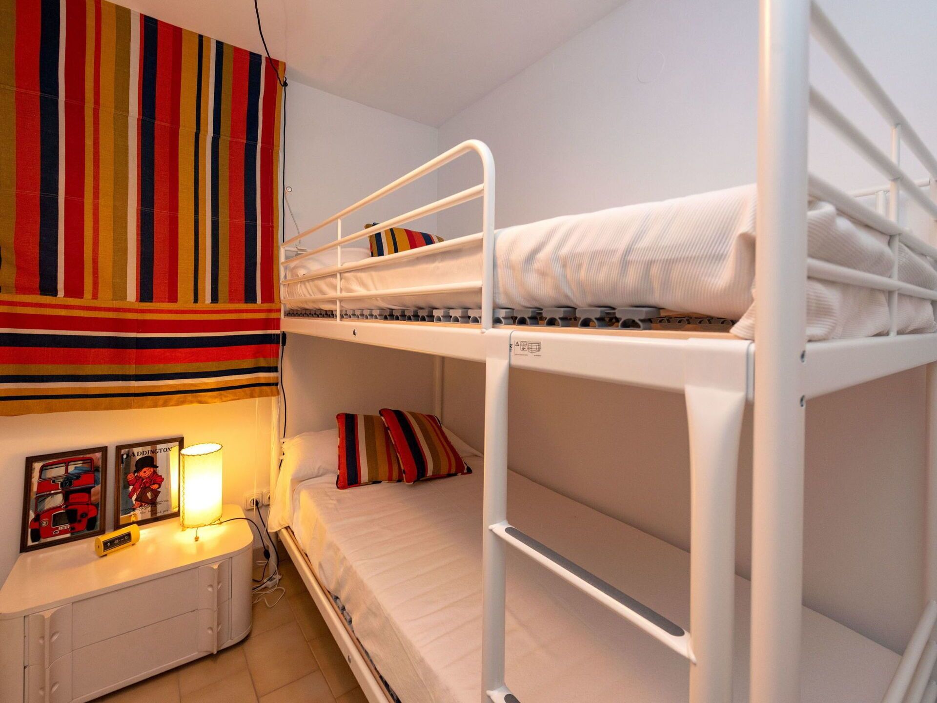 Een slaapkamer met stapelbed, een gordijn met strepen, een nachtkastje met lamp en lijstjes met foto's aan de muur.