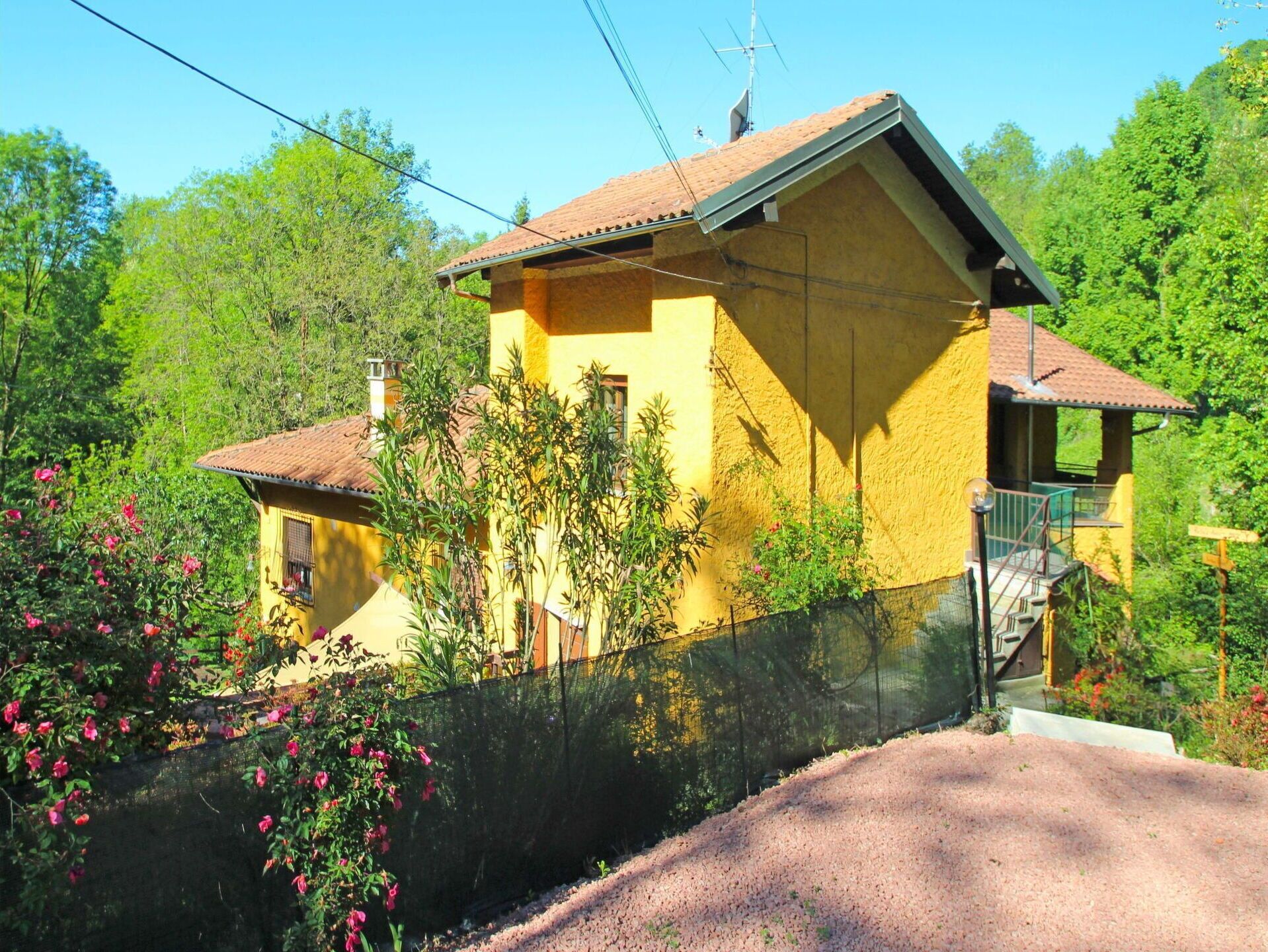 Une maison jaune avec un toit rouge, entourée de verdure et d’une clôture.