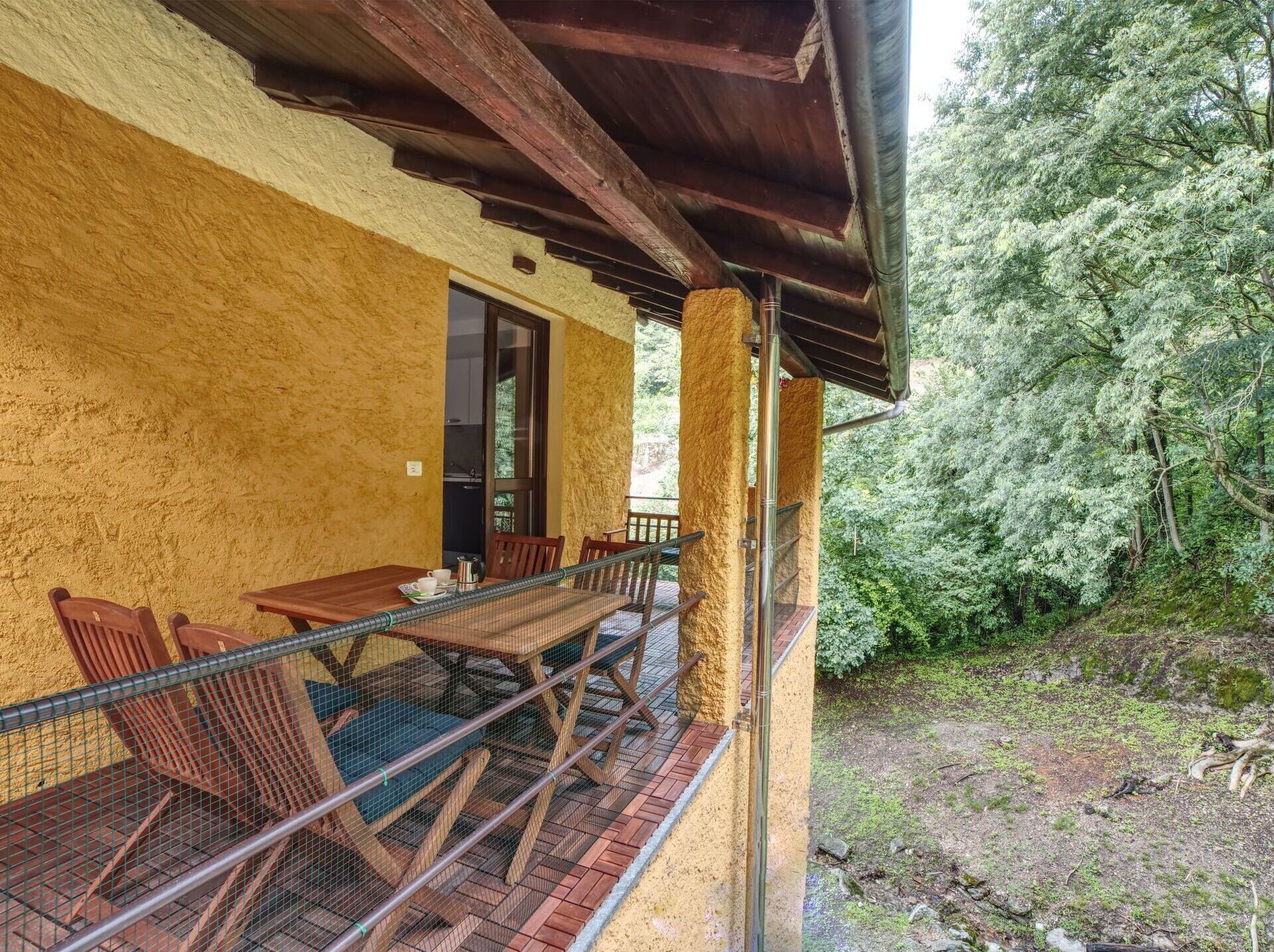 Un espace extérieur abrité, doté d’une table et de chaises en bois, d’une rambarde en métal et offrant une vue sur une zone boisée.