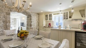 A dining area with a table set for four, a chandelier, stone walls, and a kitchen with white cabinets and a view.