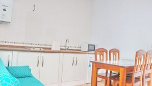 Cocina compacta con armarios blancos, un microondas y un lavabo. Hay una mesa de comedor con sillas y un sofá con cojines estampados.