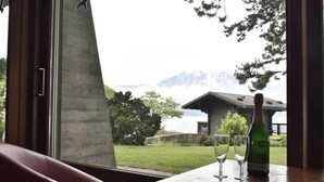 A table with two glasses and a bottle, a red bench, and a view of a mountain and a building through a window.