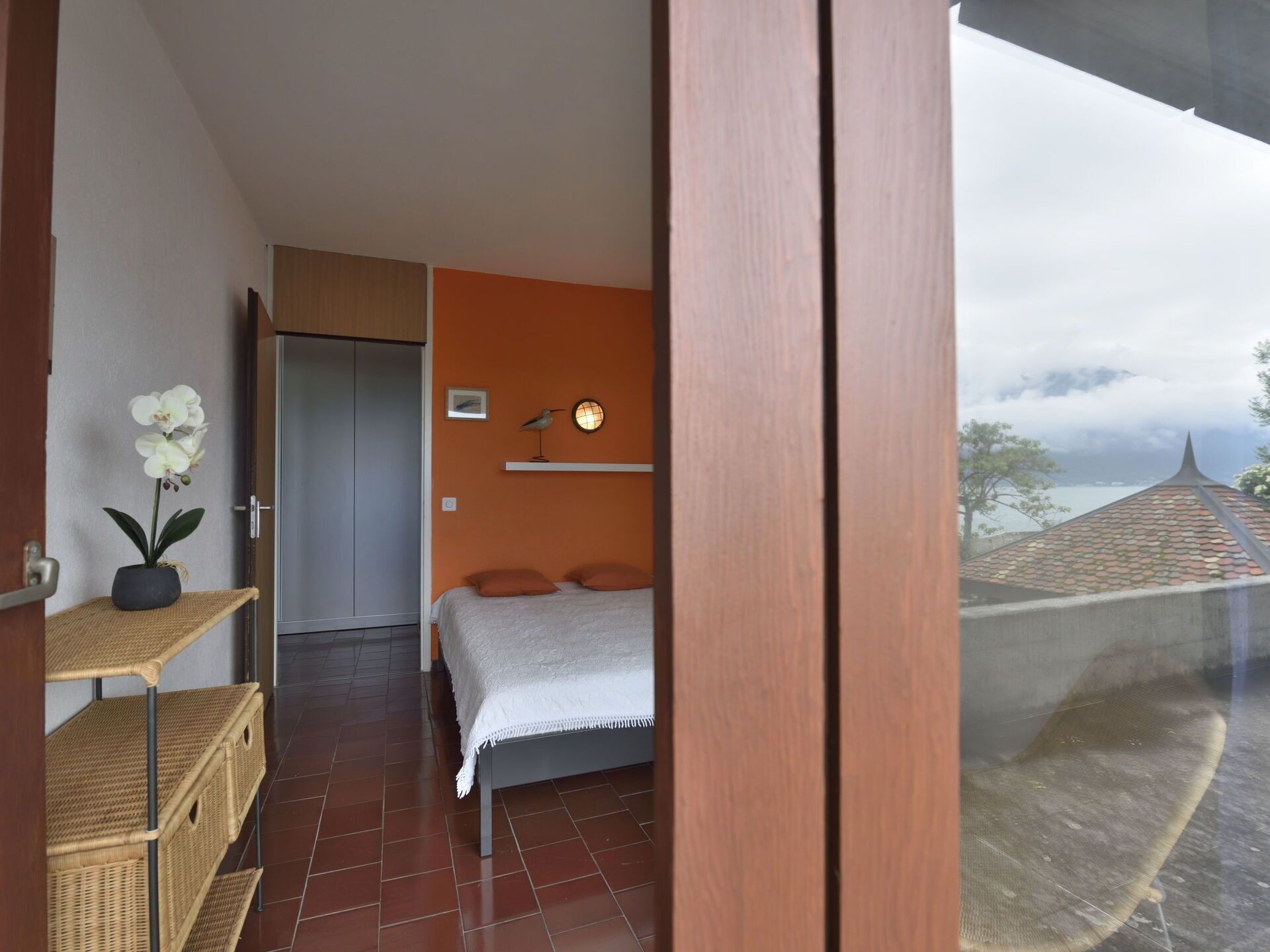 Une chambre à coucher avec un lit, une étagère ornée d’une plante en pot et une vue sur un paysage côtier.